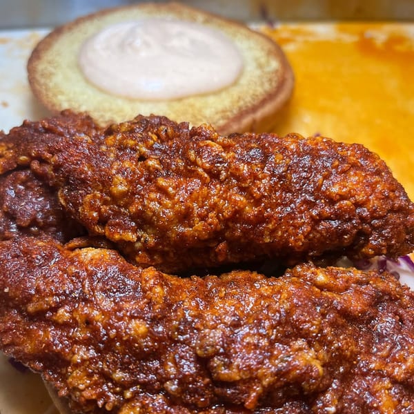 Close-up of two crispy, dark red fried chicken tenders with a bun and sauce in the background.