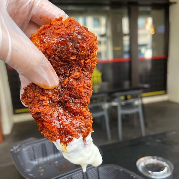 A gloved hand holds a piece of Baba's Hot Chicken, dripping with white sauce, against an outdoor background.